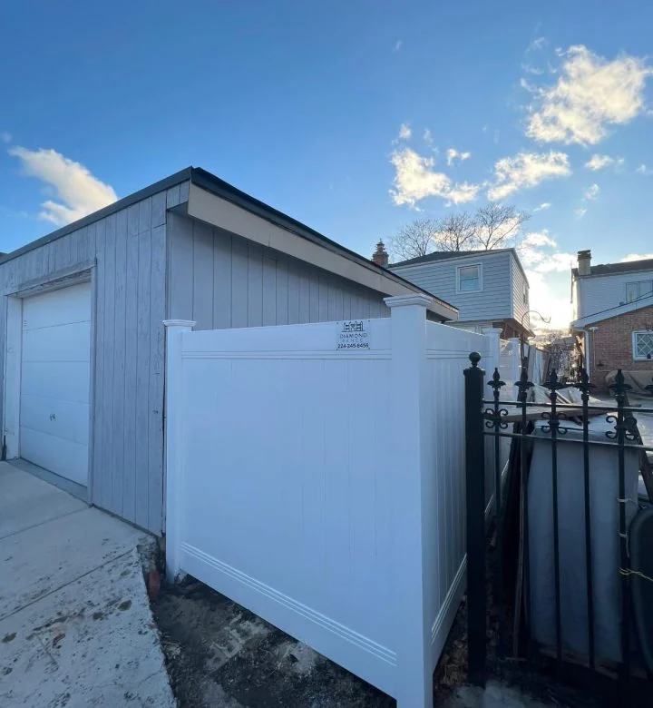 Clean white vinyl privacy fence installed beside gray garage and adjacent to black wrought iron gate, with Diamond Fence sign on corner post, perfect for narrow spaces, utility screening, or side-yard privacy