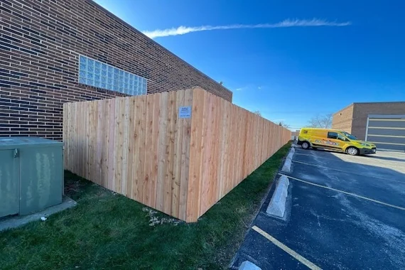 High solid wood privacy fence enclosing commercial building parking lot under clear blue sky, includes utility box concealment and branded signage