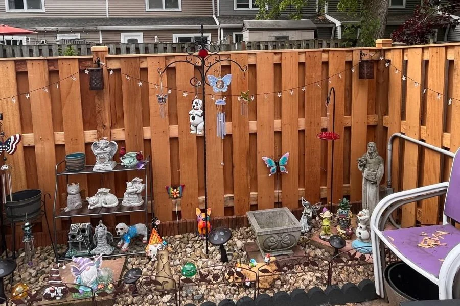 Warm-toned shadowbox wood fence backdrop for whimsical garden display with statues, wind chimes, string lights, and pebble ground cover, perfect for personal outdoor spaces
